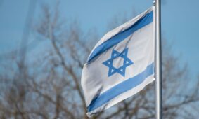The Israeli flag featuring the Star of David against a clear blue sky and trees.