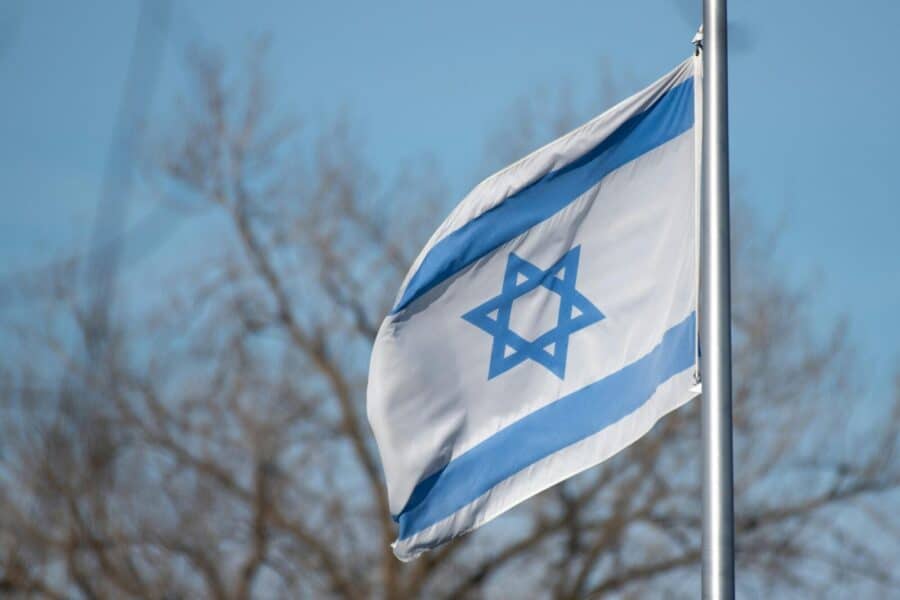 The Israeli flag featuring the Star of David against a clear blue sky and trees.