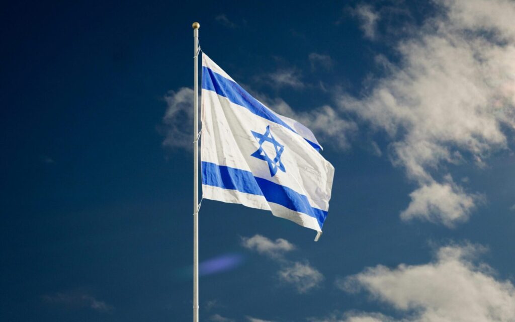 The Israeli national flag waving against a clear blue sky with clouds.