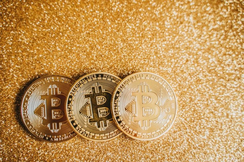 Close-up of three shiny Bitcoin coins on a sparkling gold glitter background, symbolizing cryptocurrency wealth.
