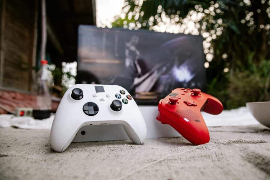 White and red Xbox wireless controllers set outdoors. A computer screen is blurred in the background.