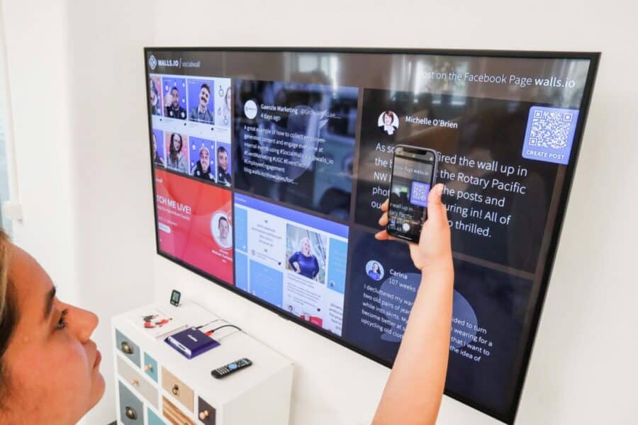 A woman uses a smartphone to scan a QR code on a digital wall display indoors.
