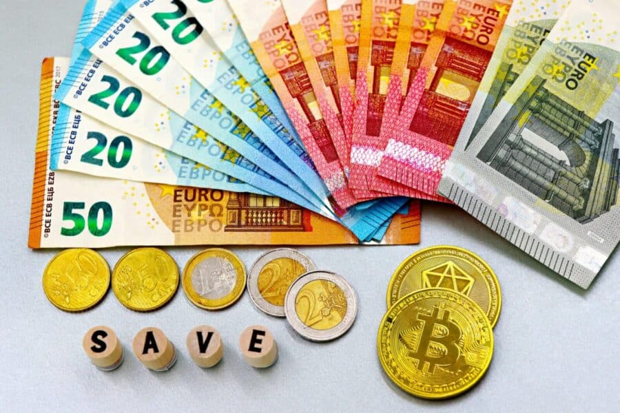 Euro banknotes and cryptocurrency coins arranged on a white surface with blocks spelling 'SAVE'.